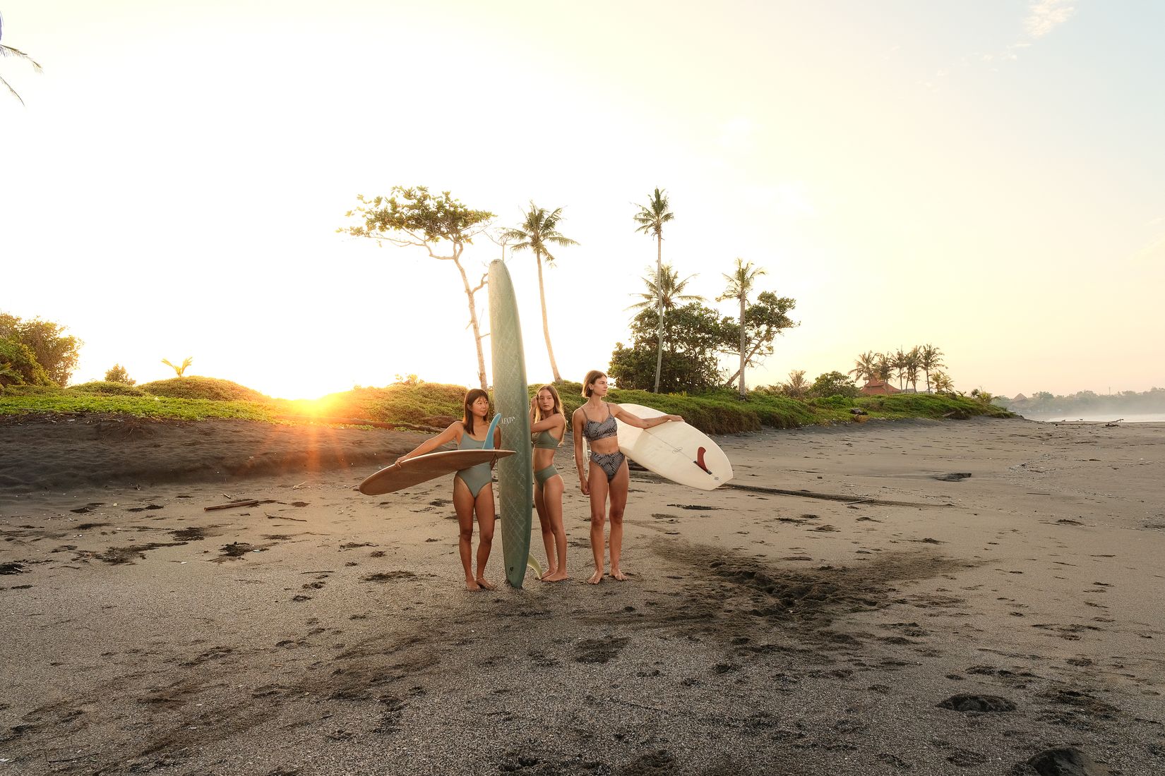 Three women holding surfboards on a dark-sand beach as the sun sets behind palm trees