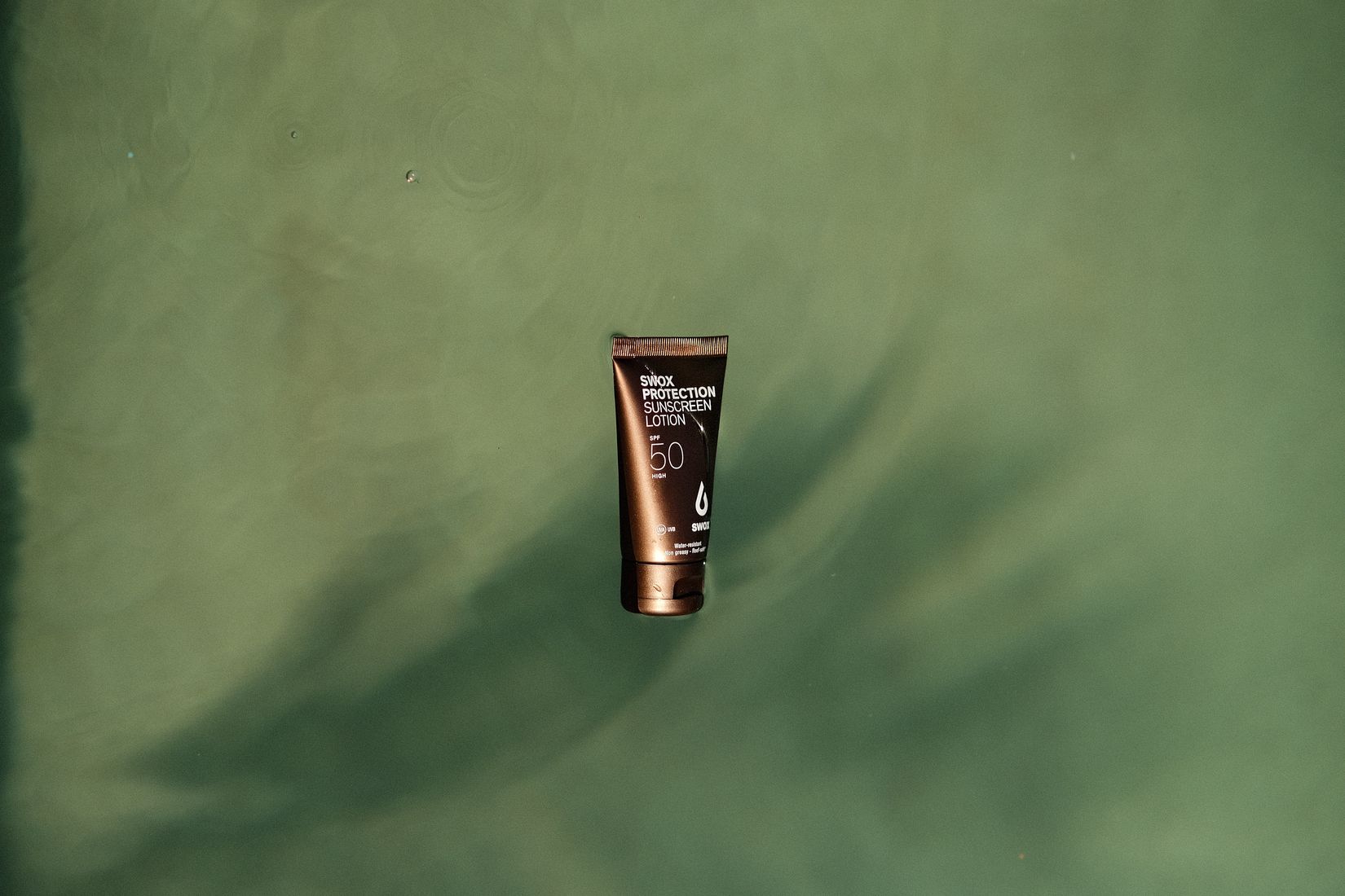 Tube of Swox SPF 50 sunscreen floating on green water with palm shadows