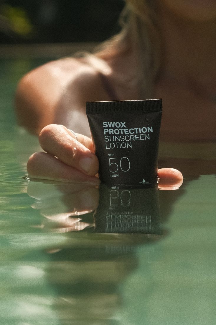 Woman in a pool holding a Swox SPF 50 tube up at the water's edge