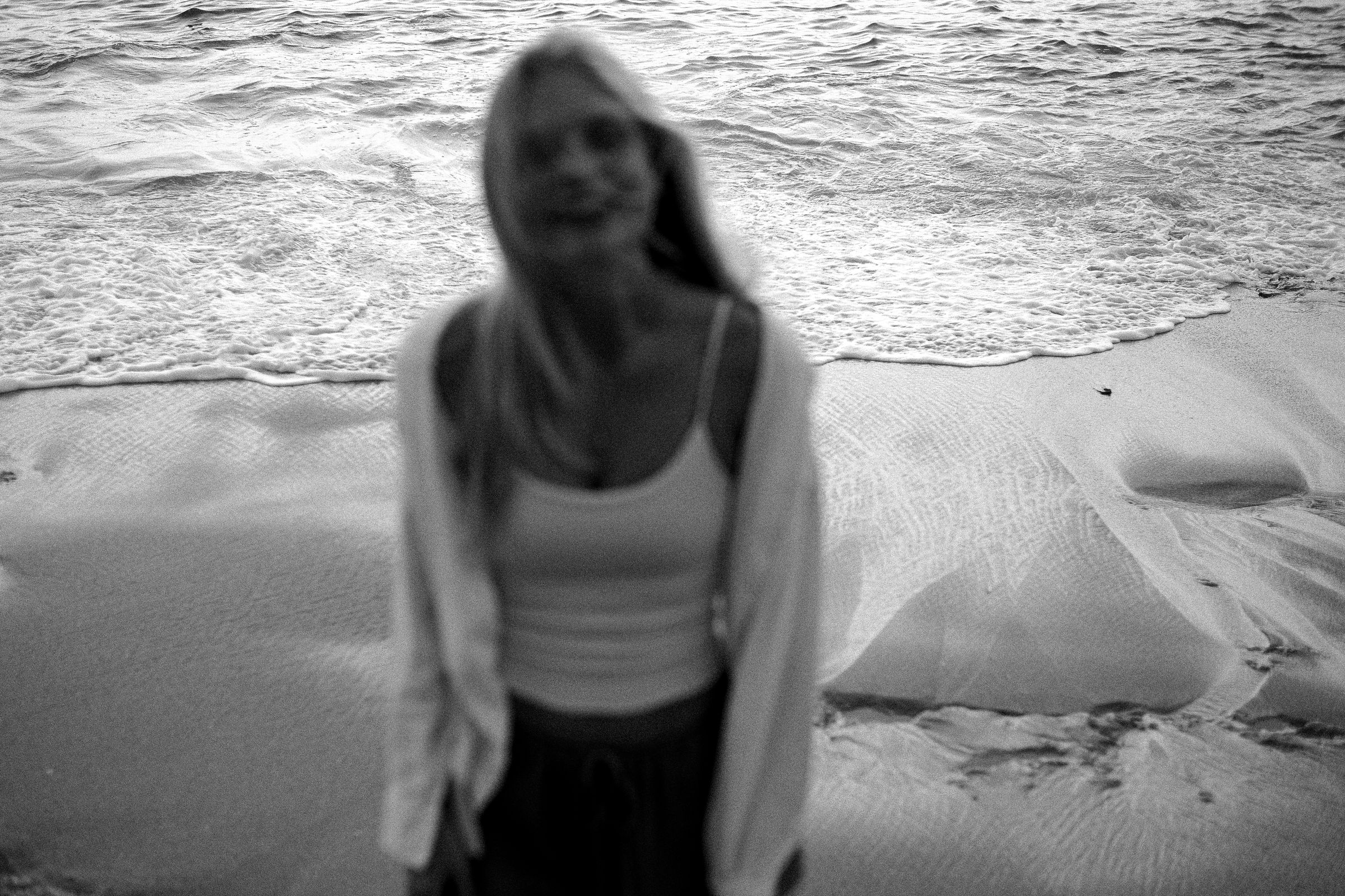 Black and white, softly blurred shot of a woman in a white top and open shirt on the beach, waves behind her