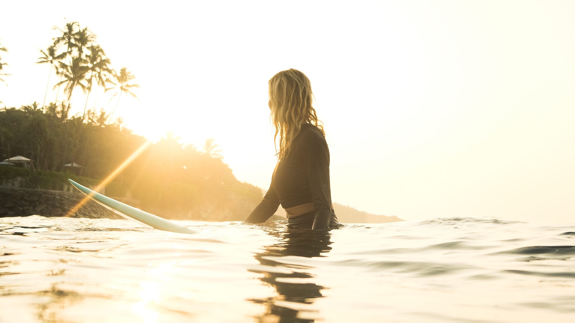 Surfer sitting in glowing golden water at sunset, palm trees behind