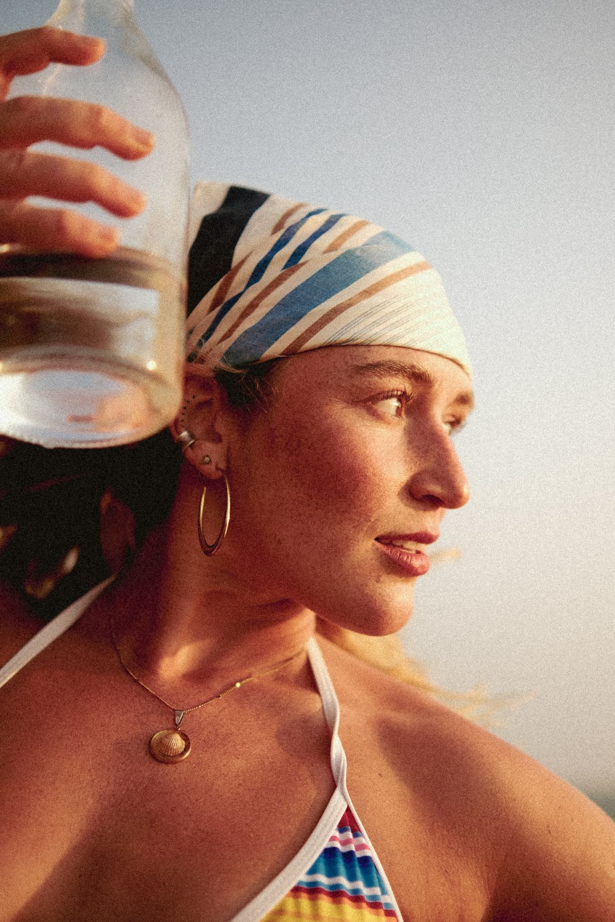 Woman in a striped headscarf holding a glass bottle to her head, gold hoops, warm light