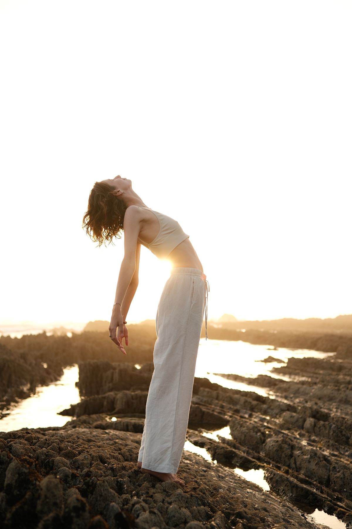 Woman in linen trousers arching backwards against the low sun on a rocky shore