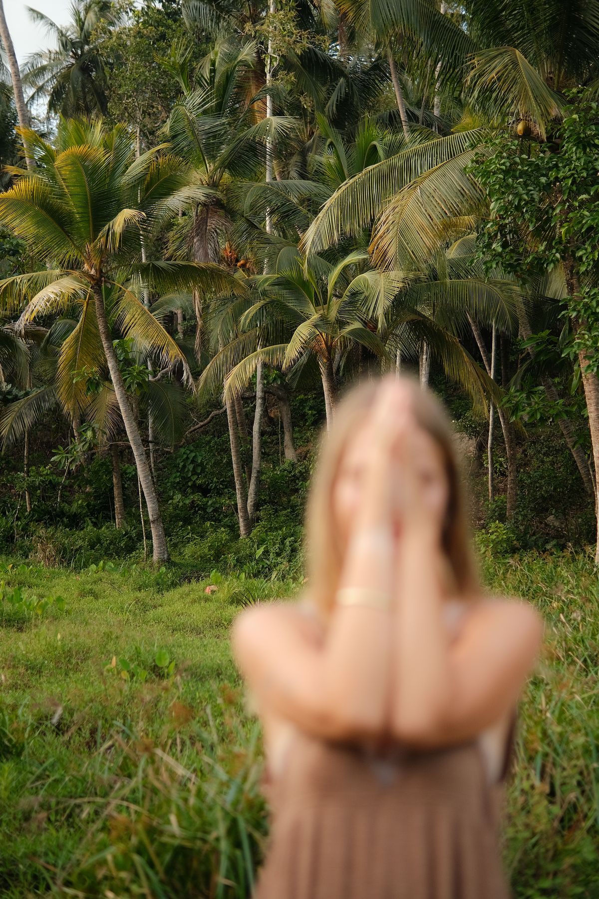 Woman with her hands over her face standing in a green field under palm trees, soft focus