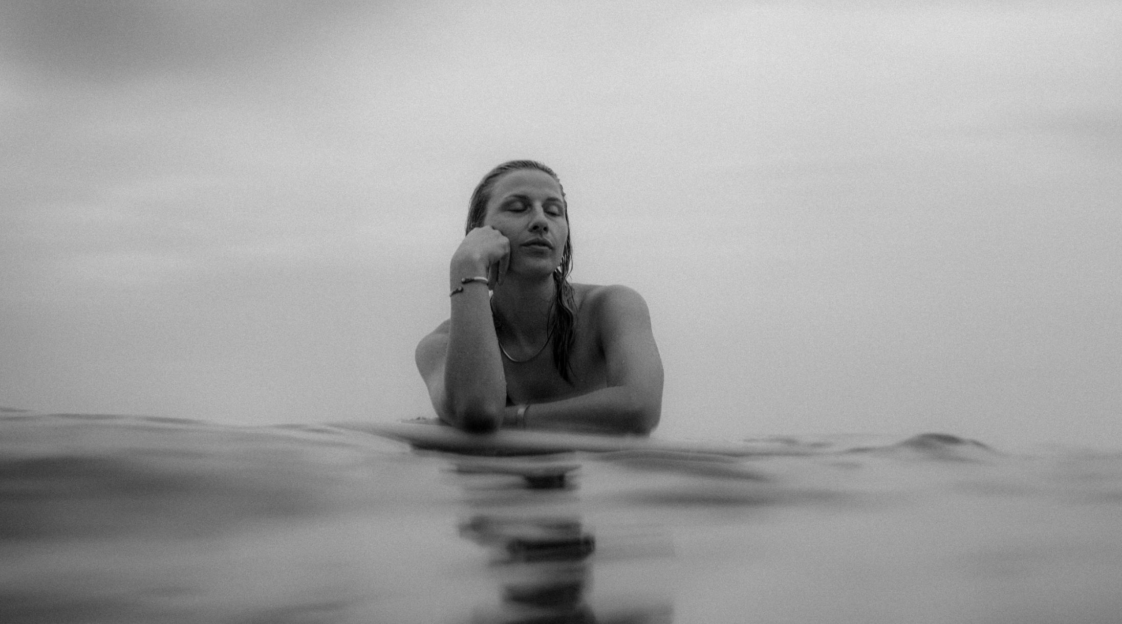 Black and white shot of a woman resting on a surfboard in calm water, eyes closed