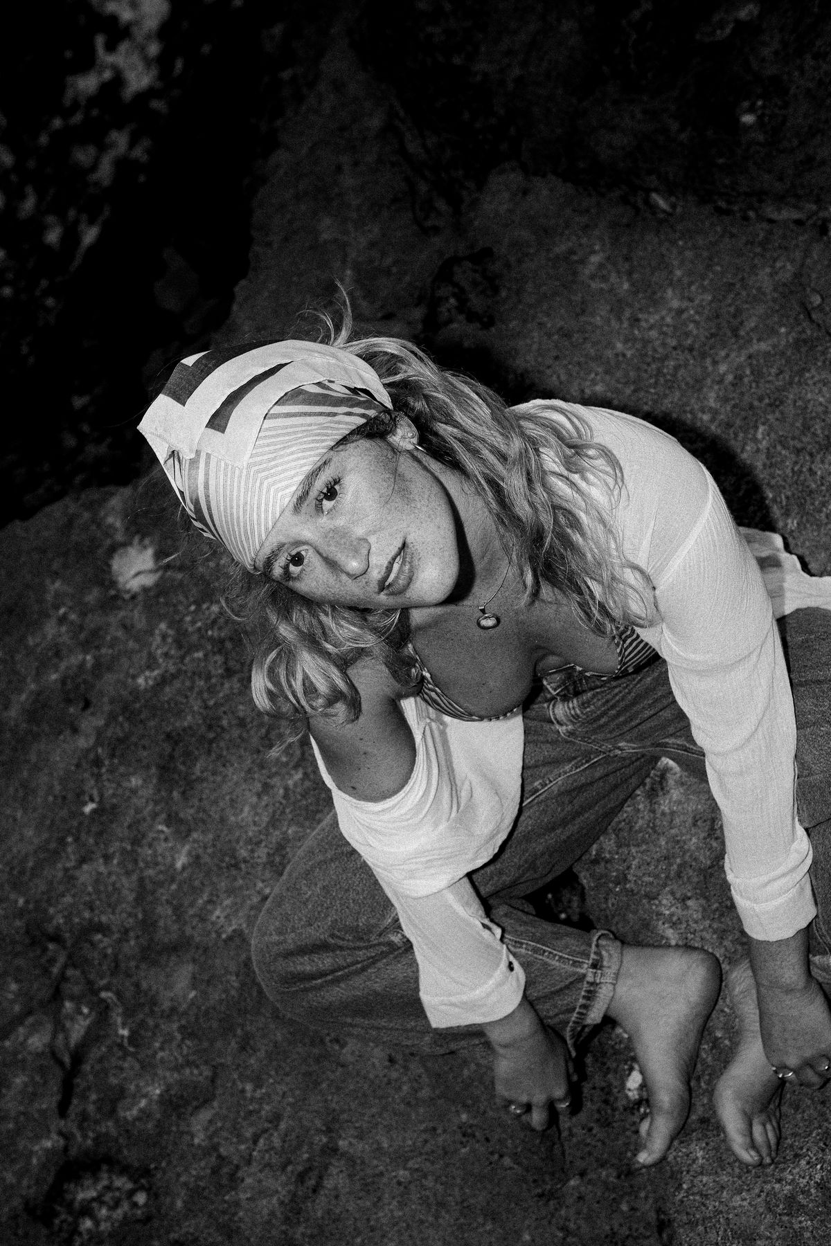 Black and white shot of a woman in a headscarf crouching on a dark rock, looking up