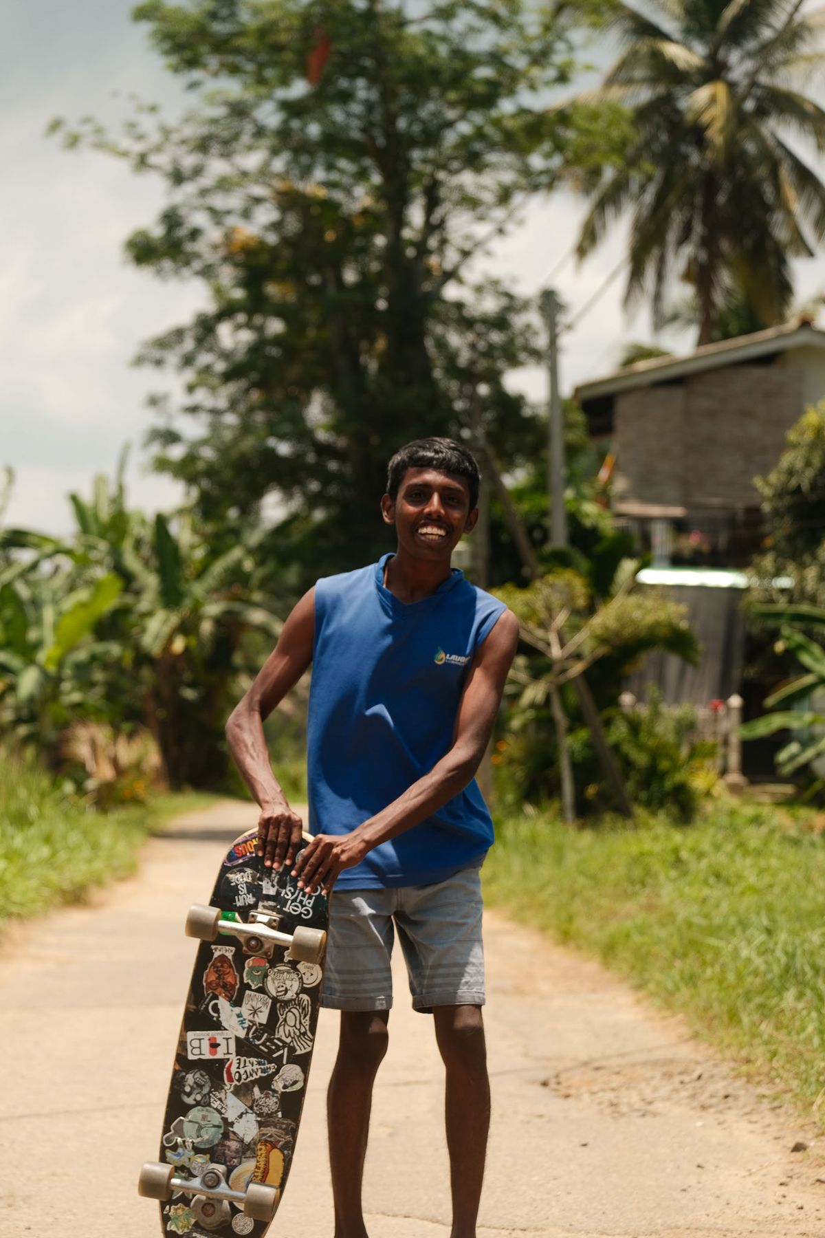 Man smiling with a sticker-covered skateboard on a jungle road