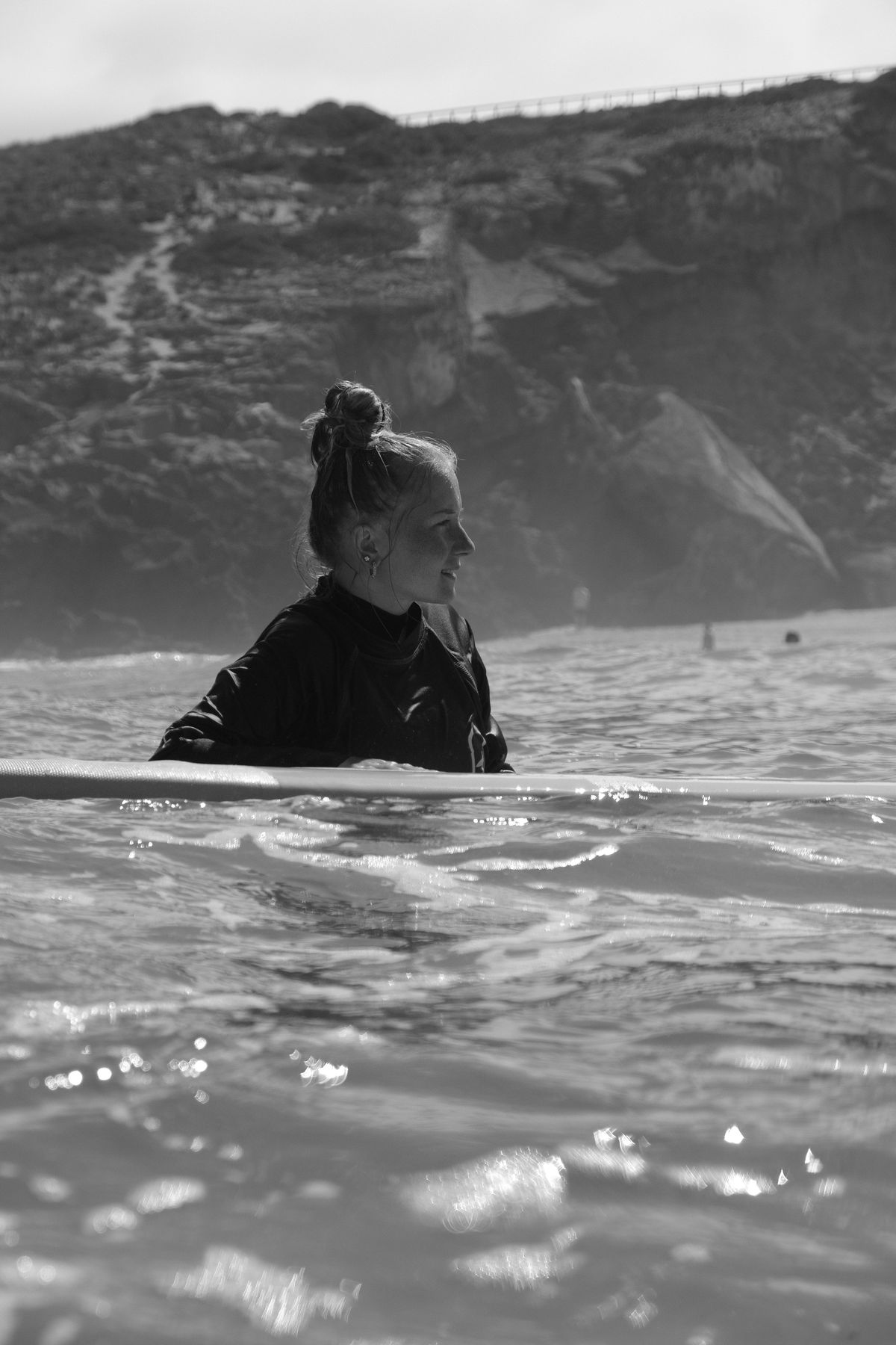 Black and white shot of a woman sitting in the water next to her board, cliffs behind