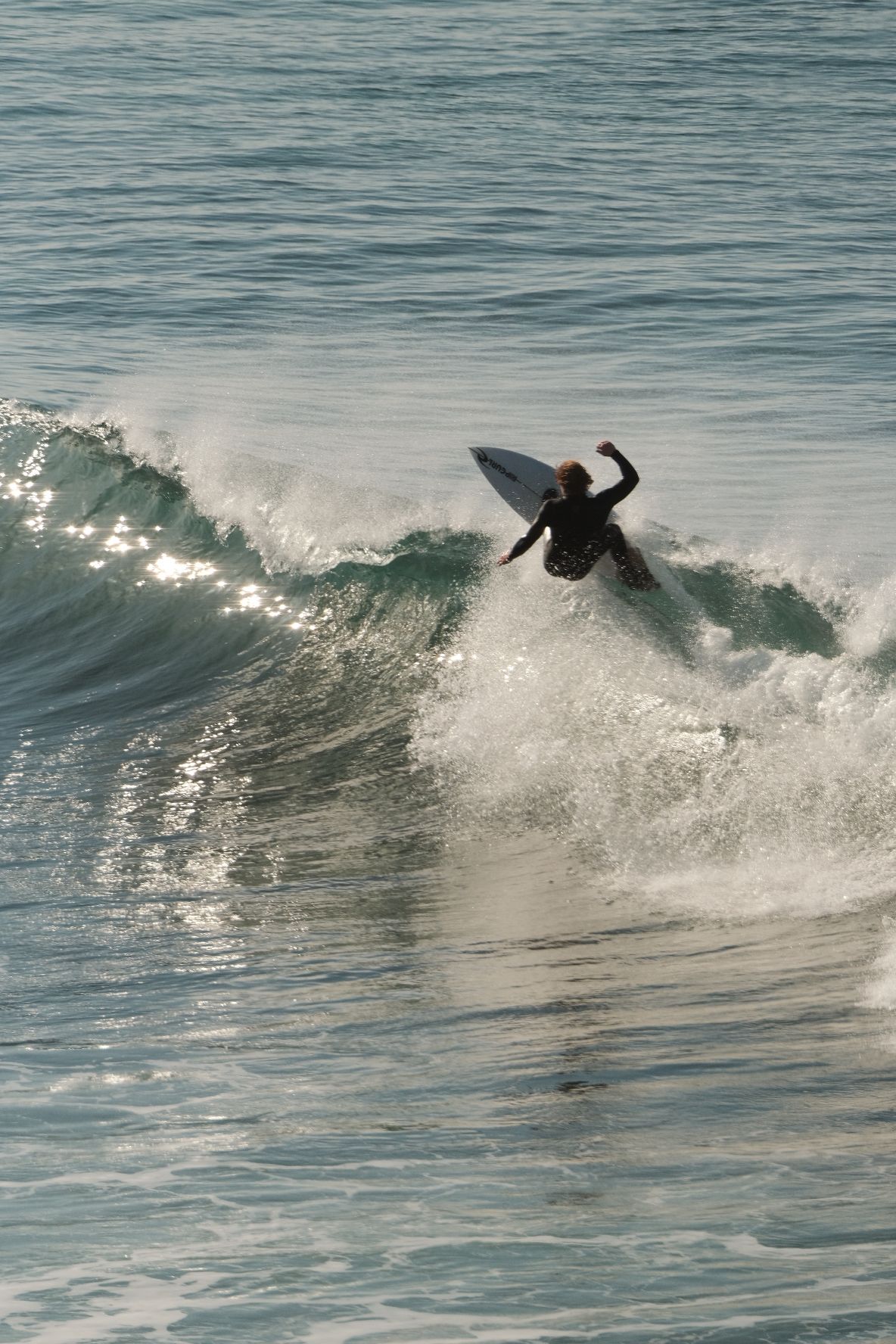 Surfer turning off the top of a wave, sun glinting on the water