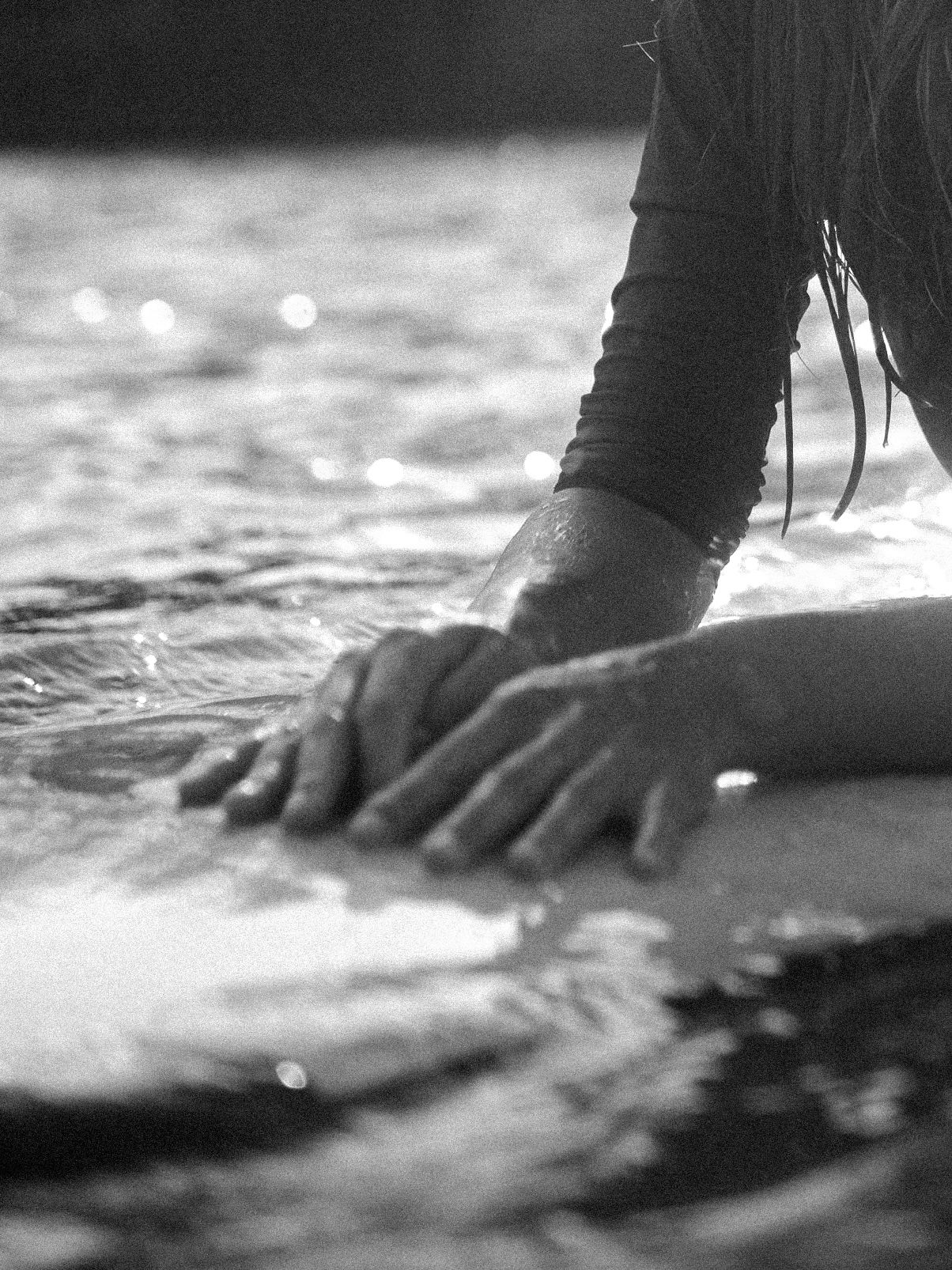 Black and white close-up of hands resting on a surfboard in the water