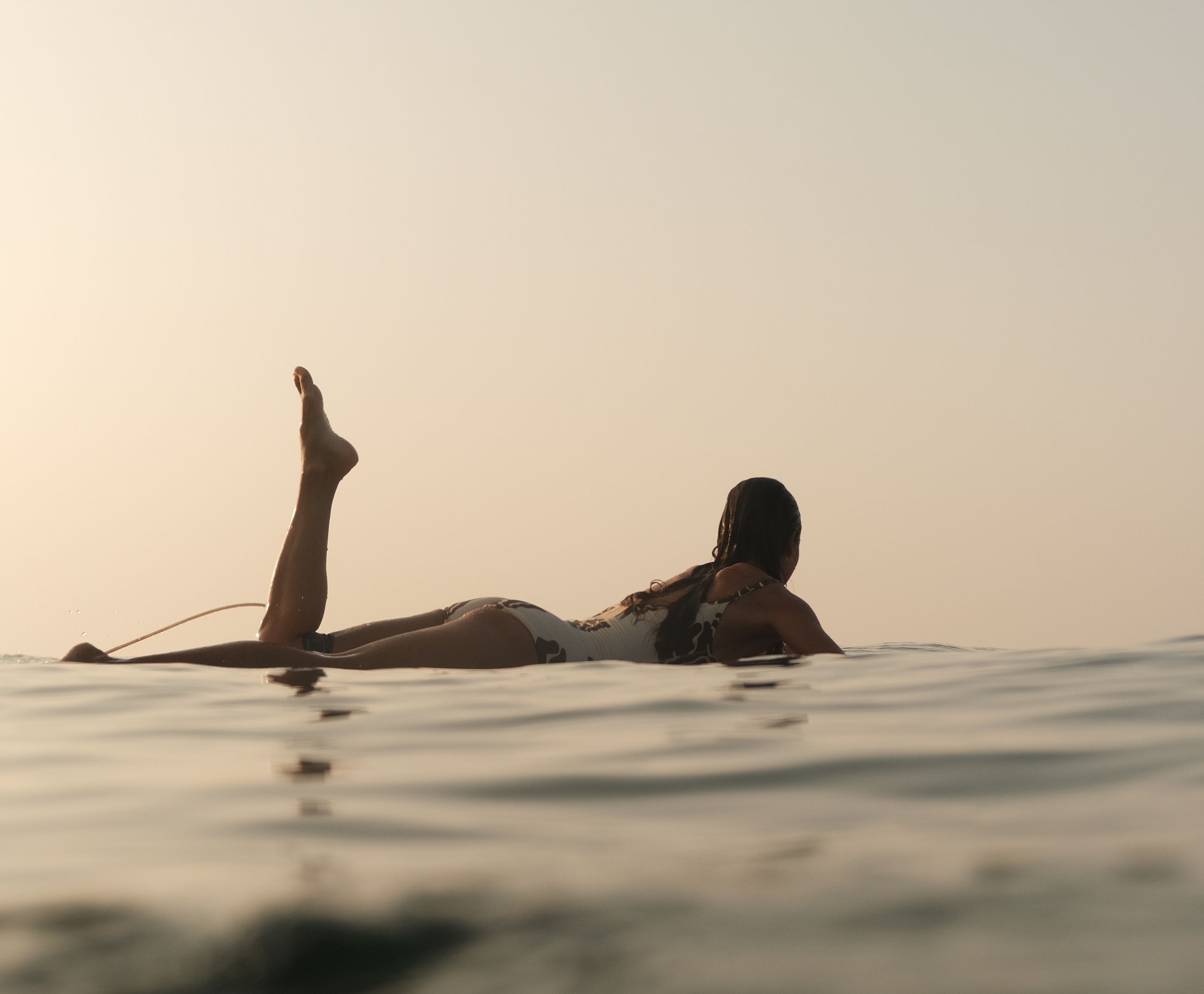 Woman lying on her surfboard in calm water at sunrise, one leg in the air
