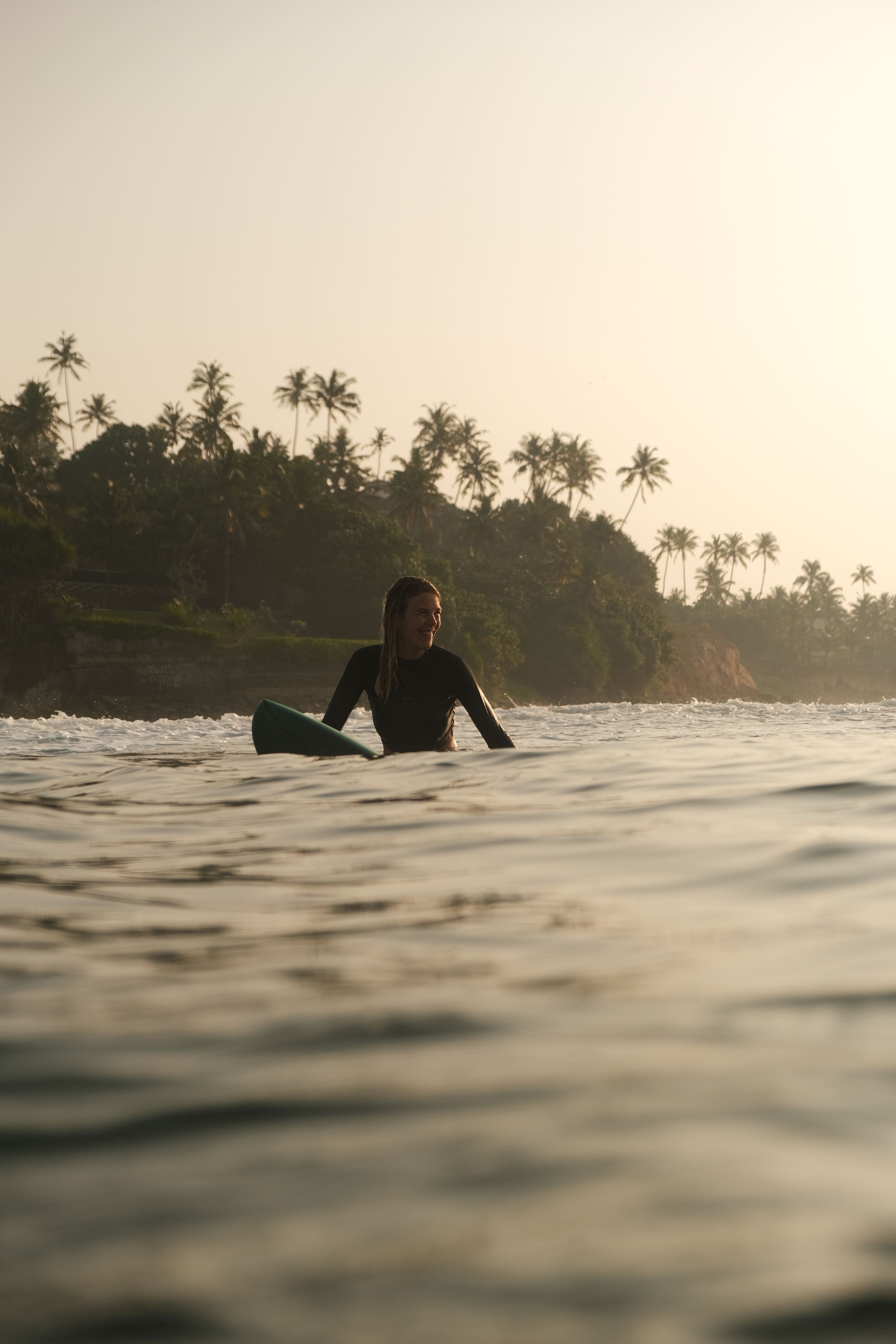 Surfer sitting on her board in calm water at sunrise, palm trees behind