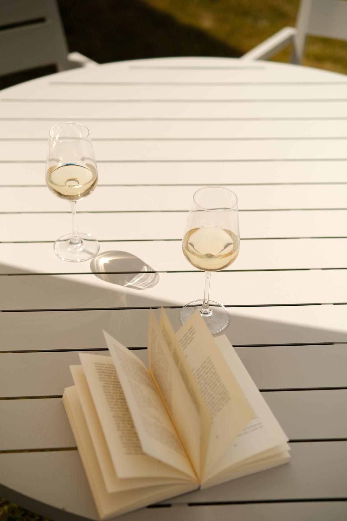 Two glasses of white wine and an open book on a white garden table
