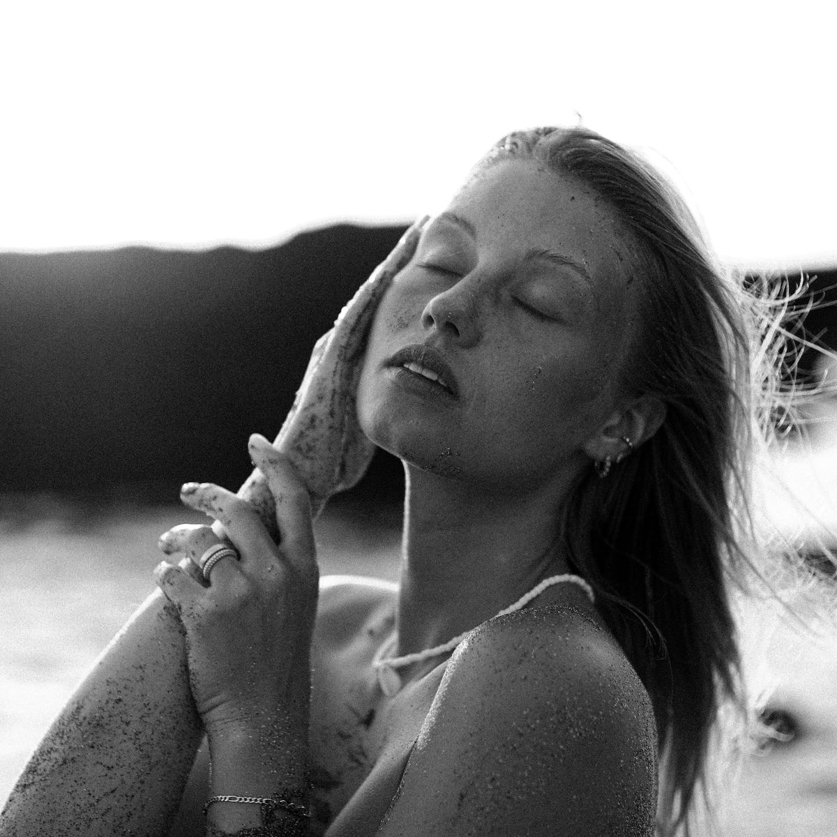 Black and white portrait of a woman holding a shell to her face, eyes closed, sand on her skin